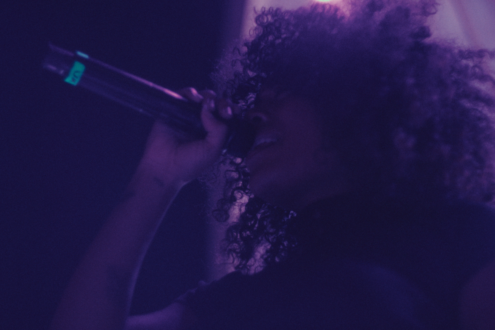 A person with curly hair holds a syringe near their face, with purple and pink lighting creating a dramatic atmosphere.
