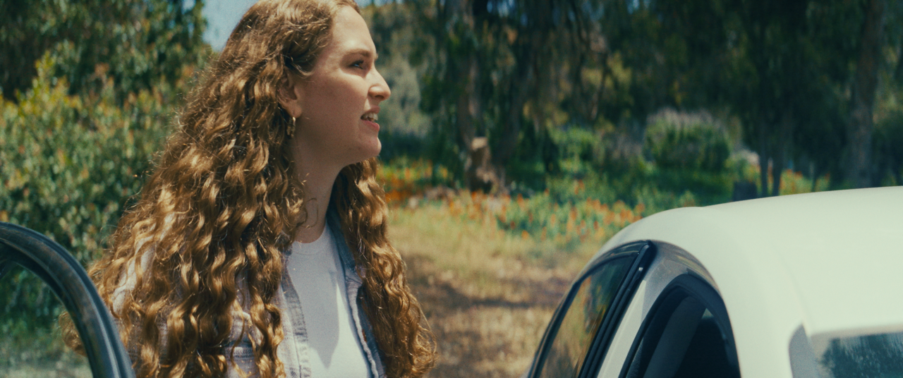 A young woman with curly hair standing outdoors near a white car and a black car, with green trees and bushes in the background.