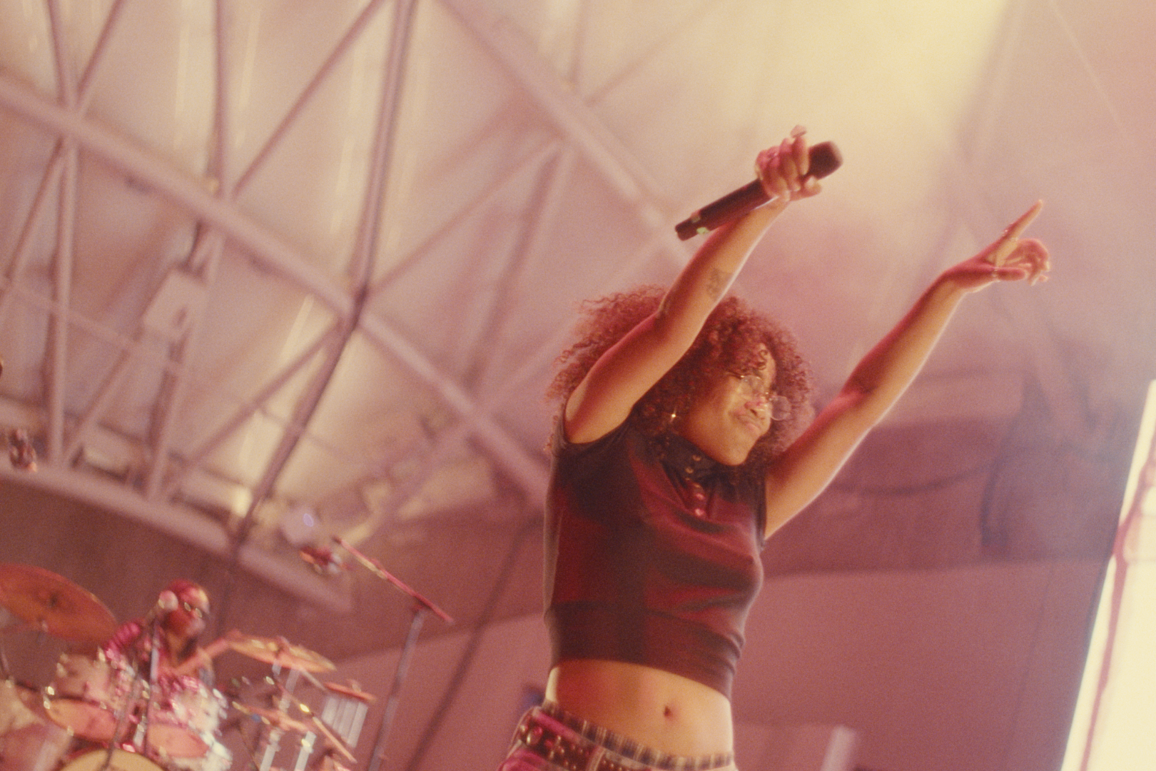 A woman with curly hair singing into a microphone on stage with a band behind her, raising her arms in the air.