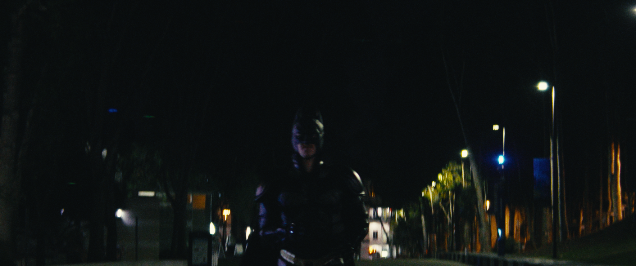 A person wearing a helmet and motorcycle gear standing at night on a street illuminated by streetlights, with trees and buildings in the background.