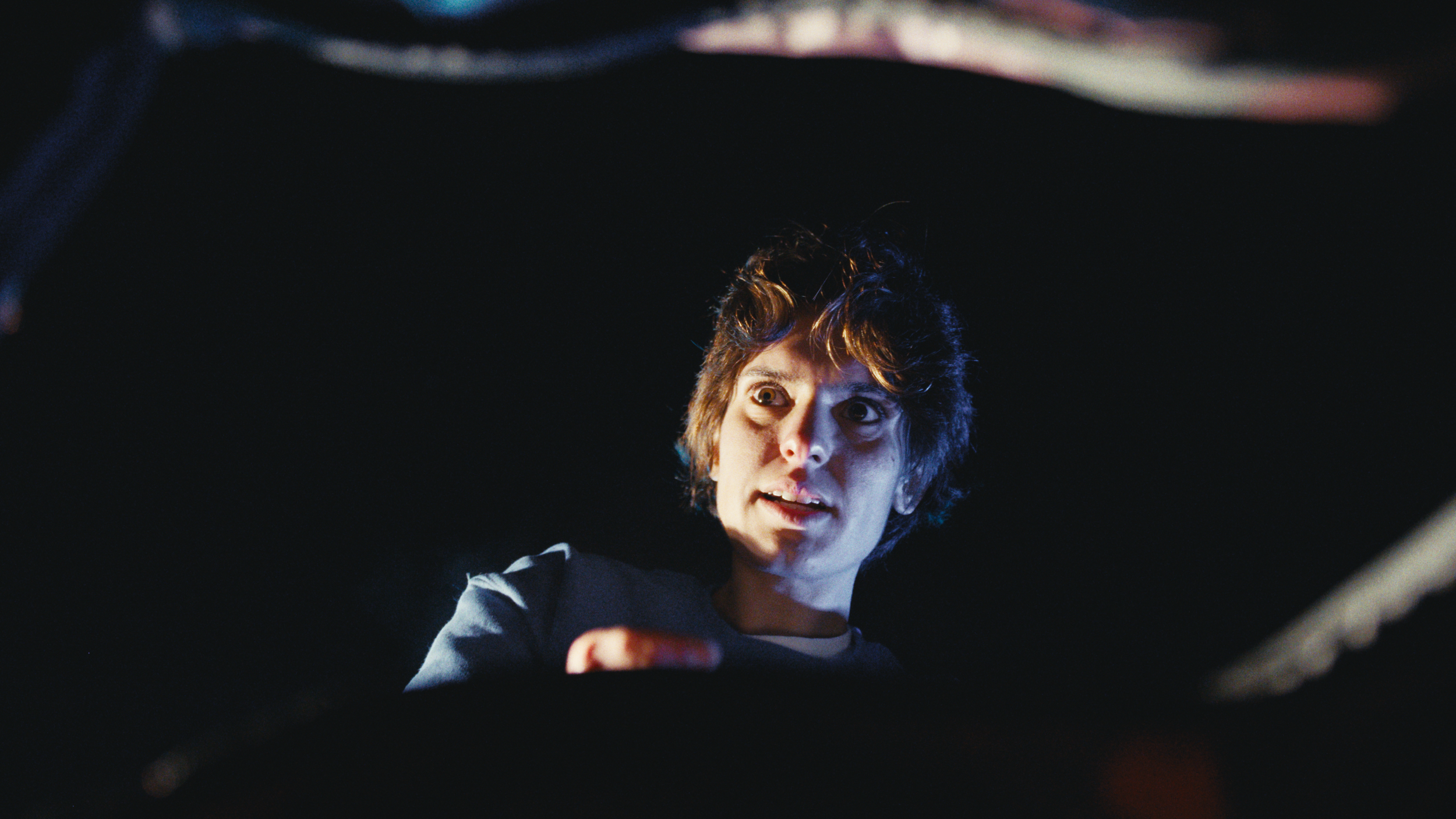 A woman with short, curly hair looks up with a surprised or concerned expression while reaching out with her arm. The background is dark and blurred.