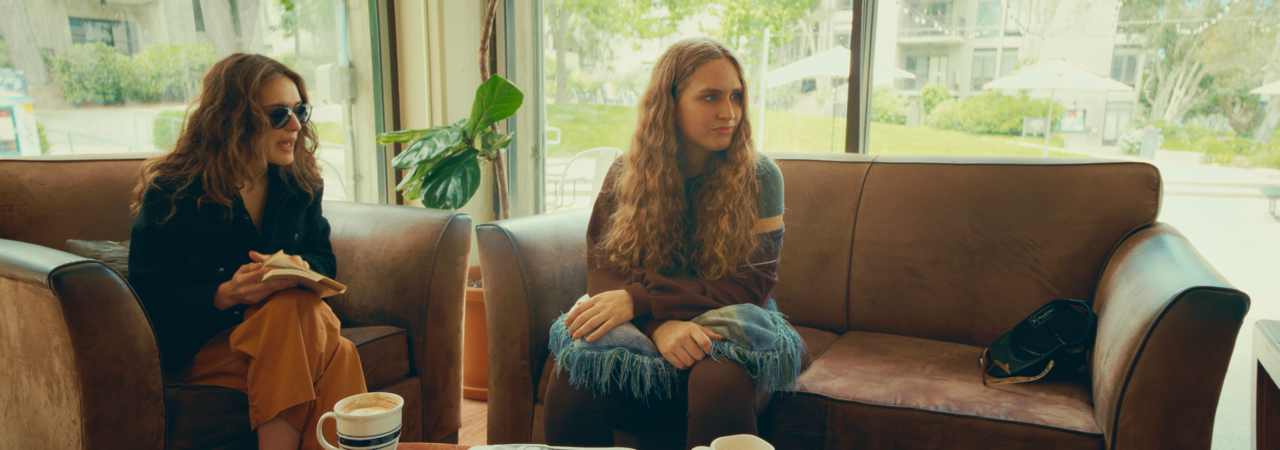 Two women sitting on leather couches in a cafe, engaging in conversation. The woman on the left wears sunglasses, a black jacket, and orange pants, holding a notebook and pen. The woman on the right has long, curly hair, and wears a multicolored swea