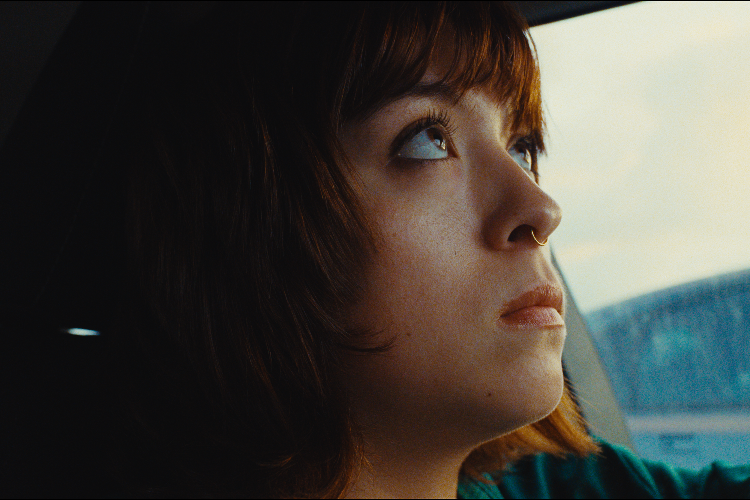 A woman with reddish-brown hair gazes up while sitting in a car.