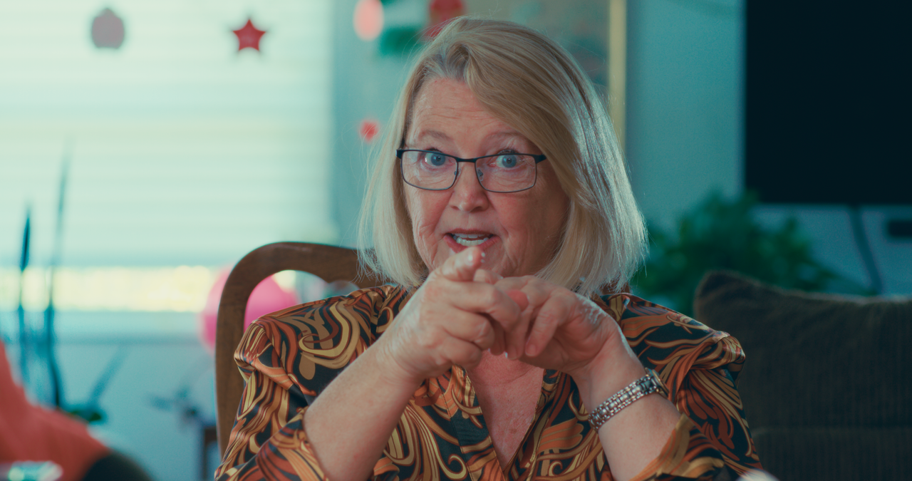 An older woman with blonde hair and glasses gesturing with her hands while sitting indoors, with holiday decorations visible in the background.