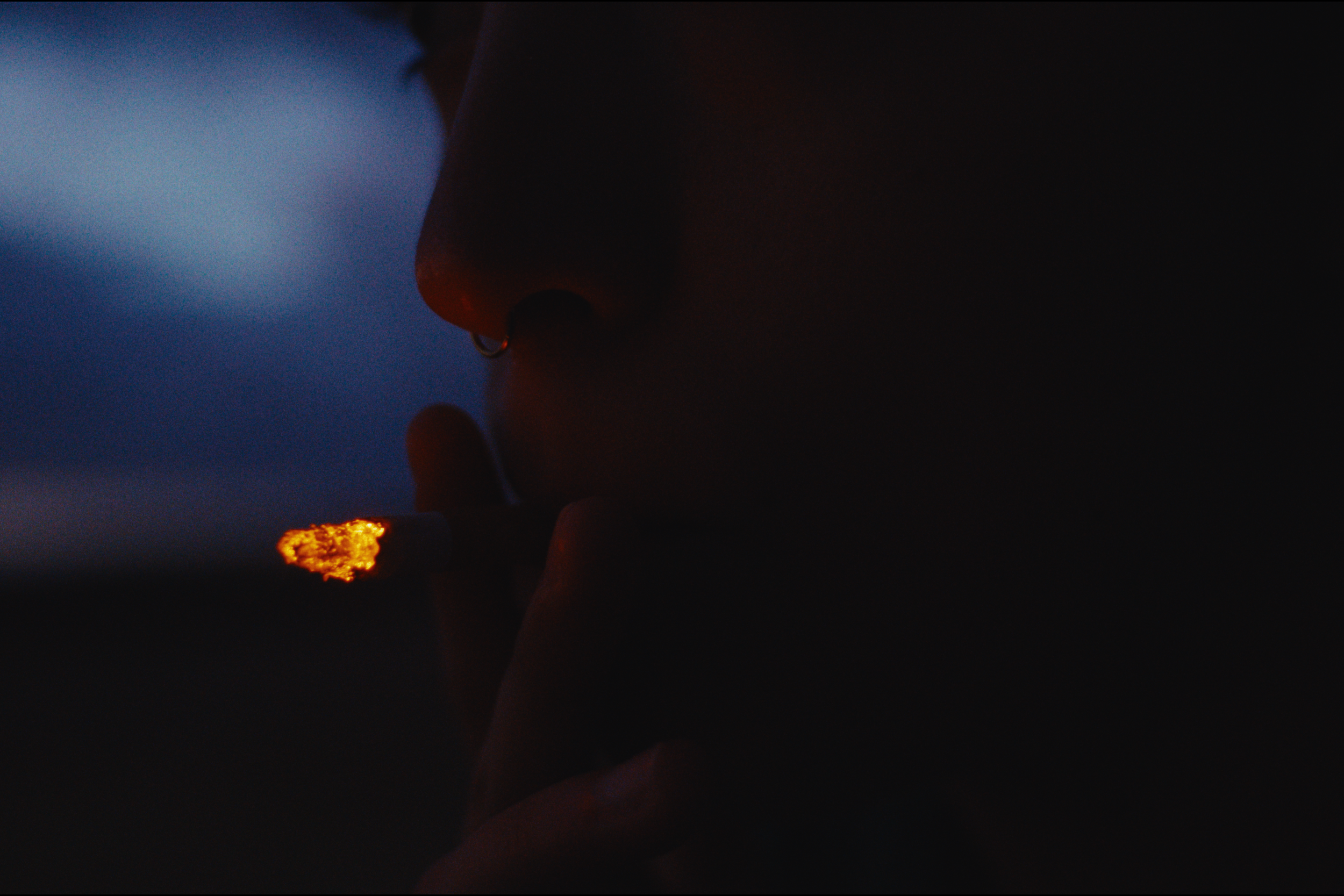 Close-up of a person's face in darkness, with a small glow from a lit object near their nose, showing a nose piercing.