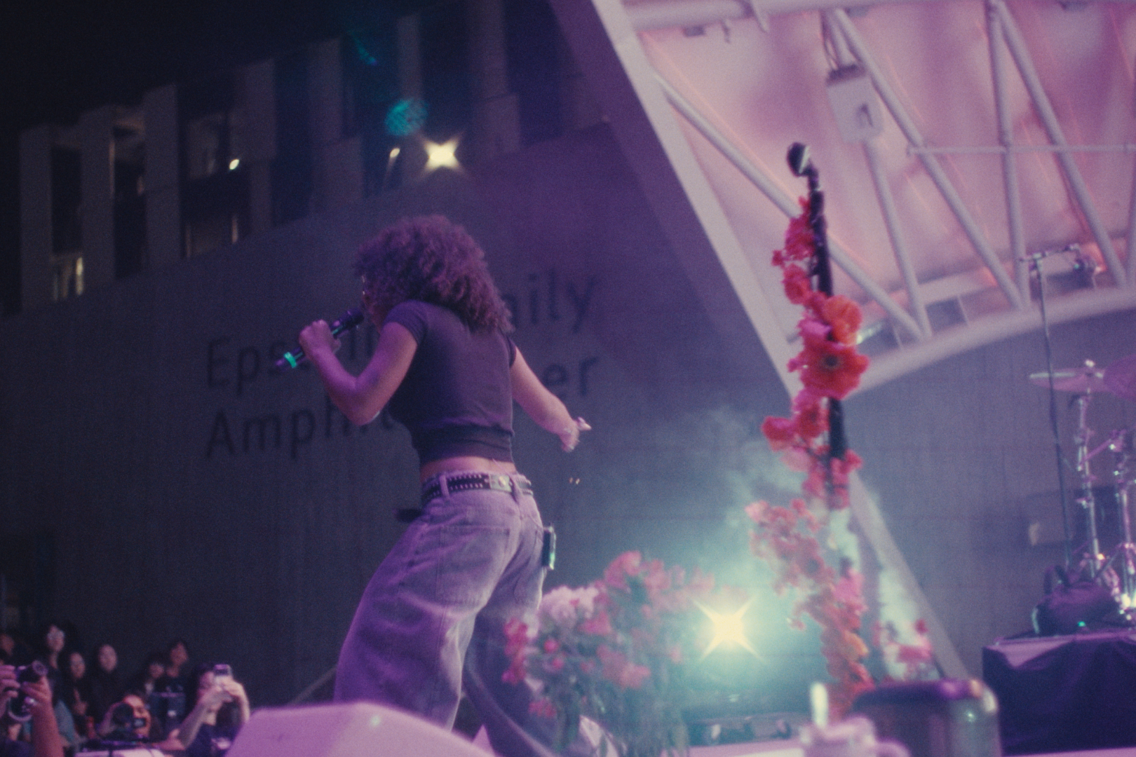 A performer singing on stage with a microphone, curly hair, wearing a black crop top and loose pants, decorated with pink flowers, in a large concert venue with audience members taking photos.
