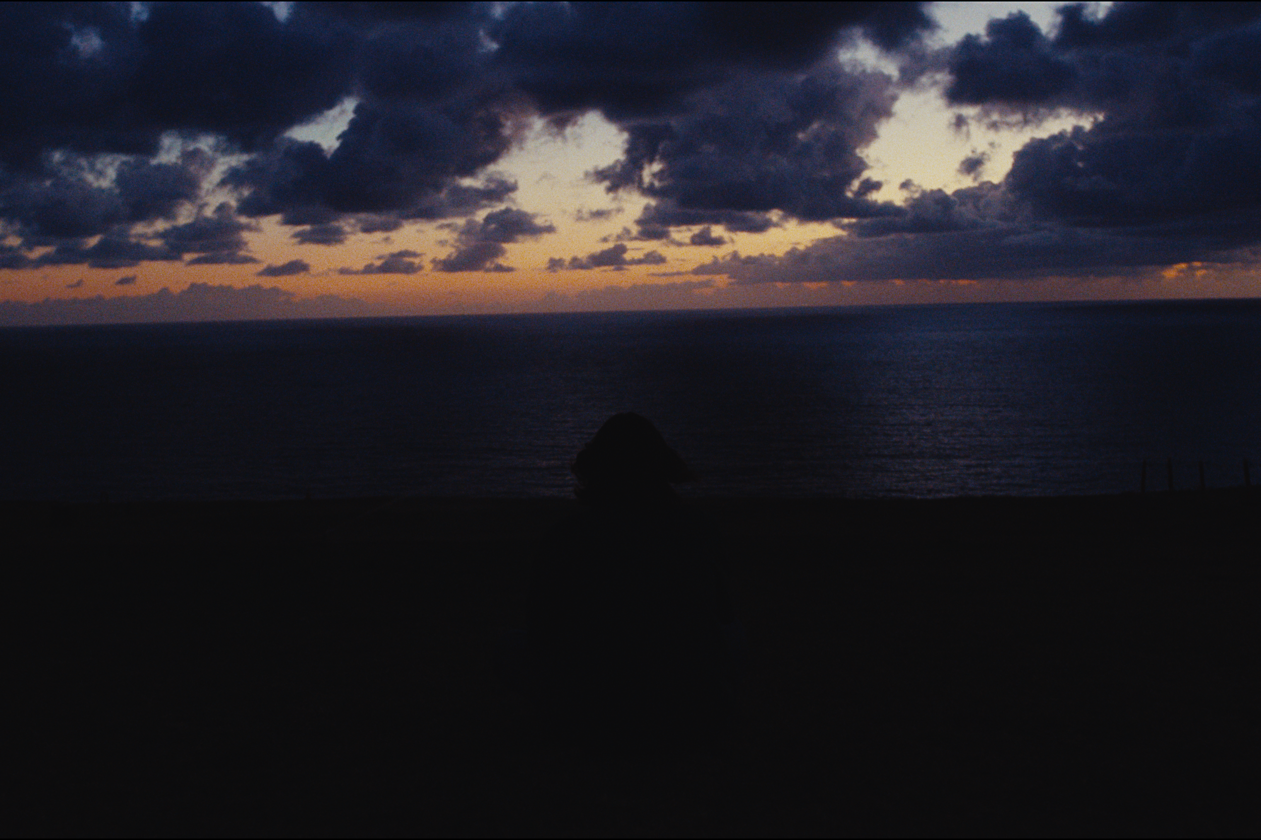 A view of the ocean during sunset or dusk with cloudy skies, silhouette of a rock formation, and a hint of orange and pink in the sky near the horizon.
