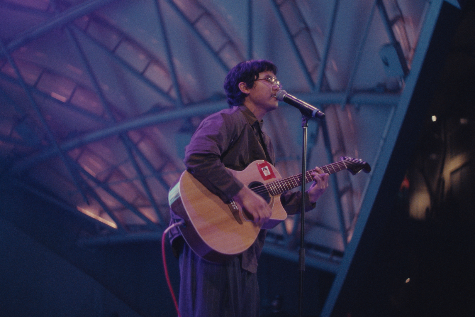 A person with glasses playing an acoustic guitar and singing into a microphone on stage. The stage has a modern, geometric ceiling design with purple and blue lighting.