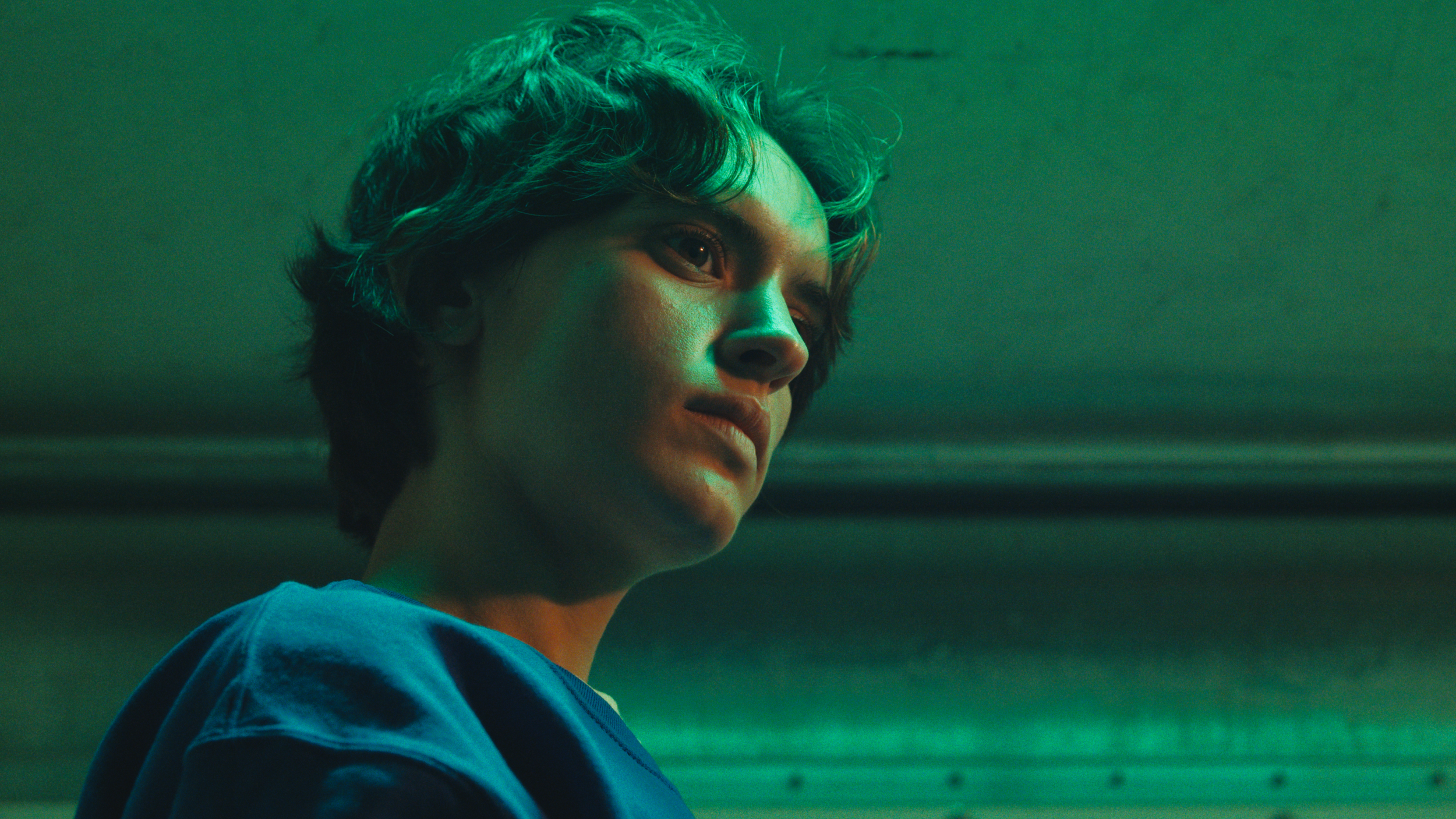 Close-up of a young person with short, curly hair, wearing a blue shirt, looking sideways with a serious expression, illuminated by greenish lighting.