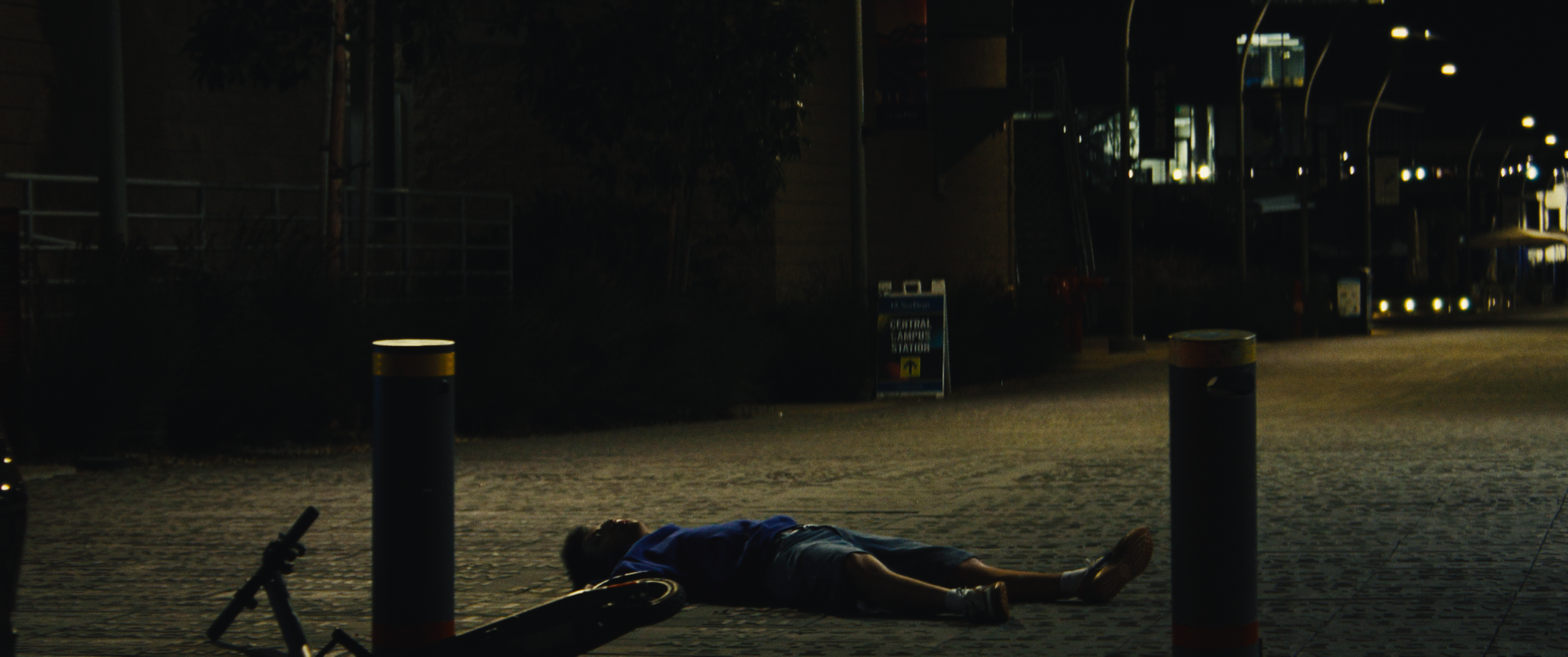 A person lying on the ground in an urban setting at night, surrounded by bollards and a bicycle, with a dark background and illuminated office buildings.