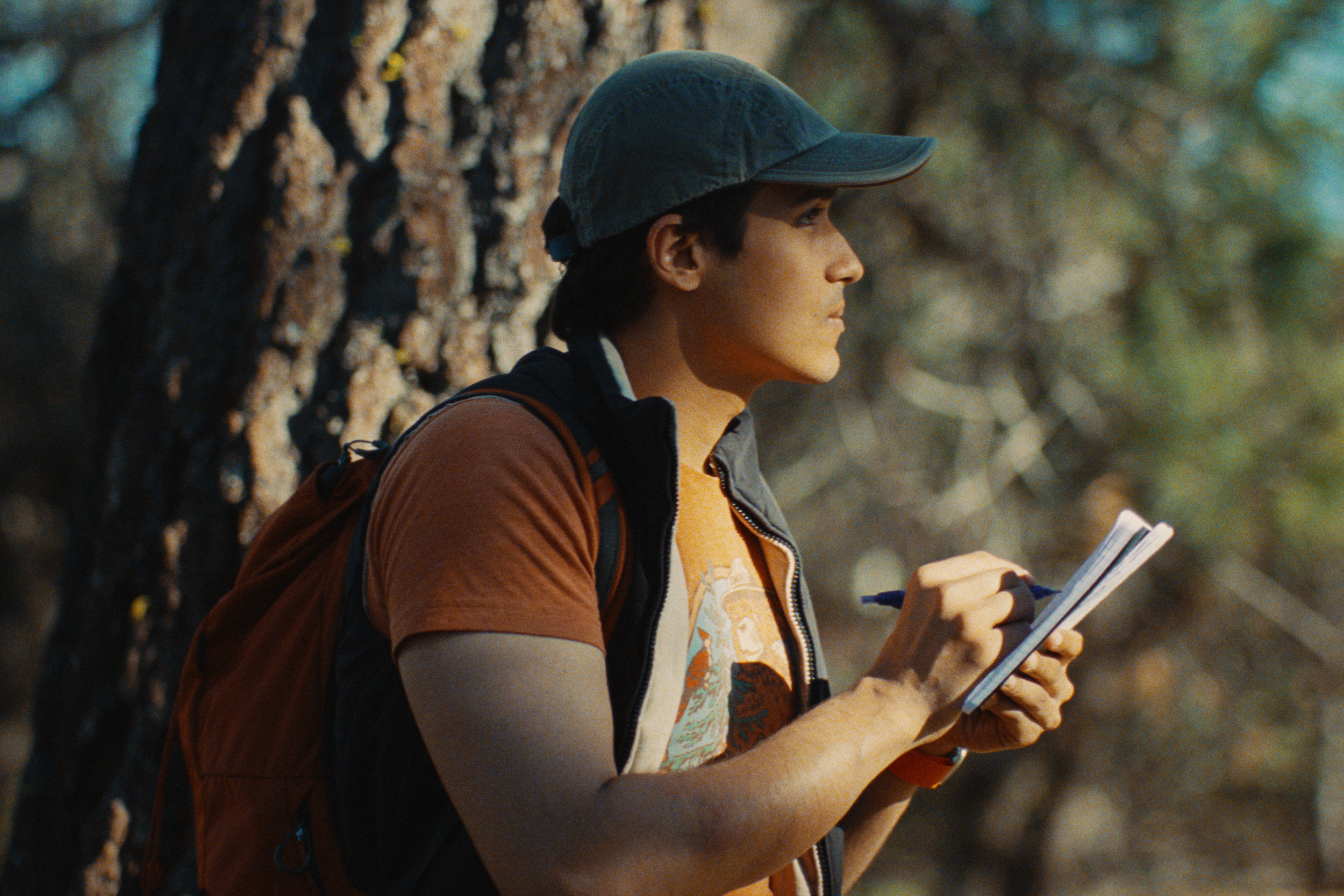 Young woman with short dark hair, wearing a baseball cap, brown jacket, and backpack, standing outdoors near a tree, writing in a notebook with a pen, holding a paper in her left hand.