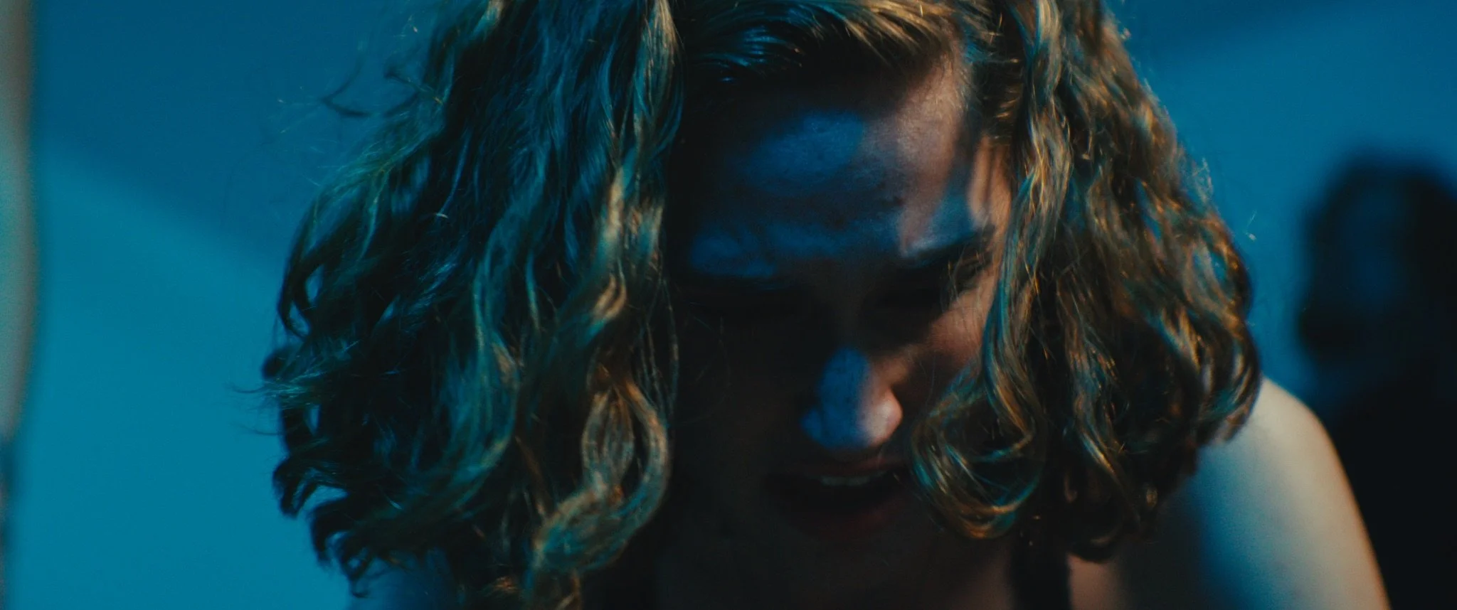 A woman with curly hair looking down, with a serious or intense expression, illuminated by blue lighting.