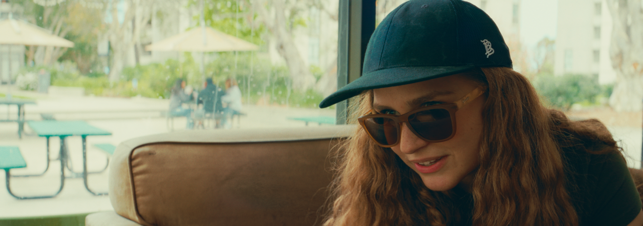 A woman with long curly red hair wearing a baseball cap and sunglasses sitting inside a cafe near a window.