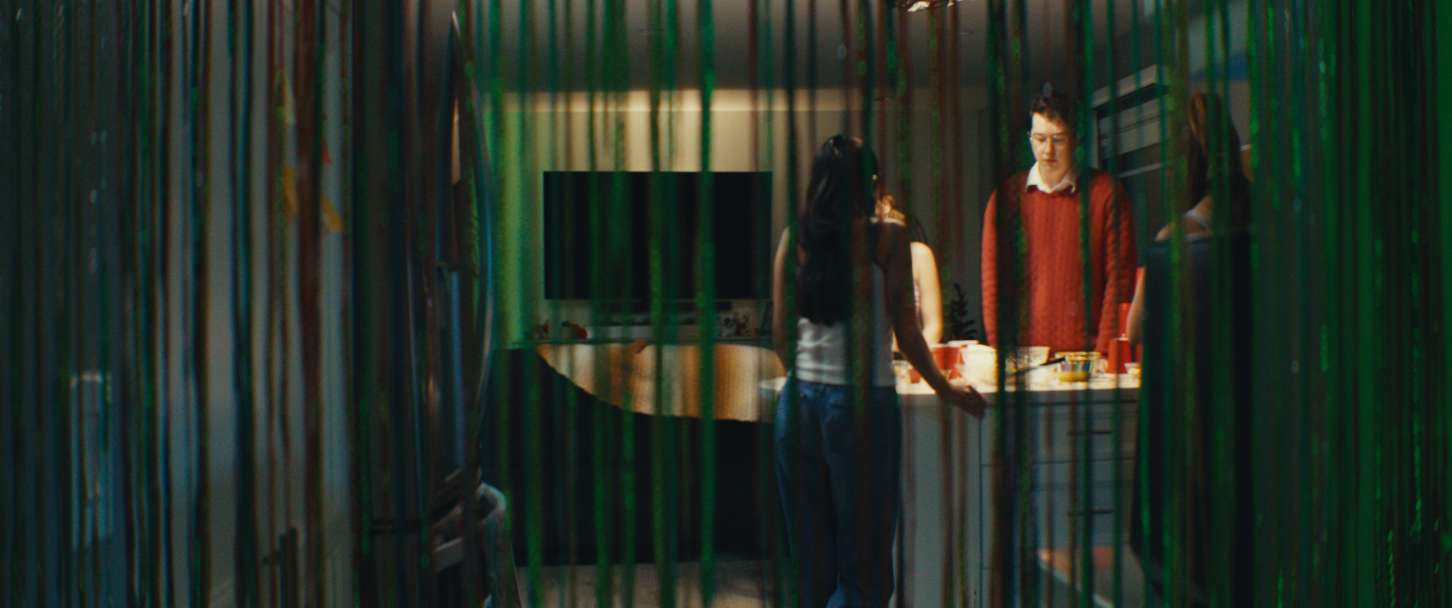 Three people seen through green beaded curtains, gathered around a table with food and drinks in a dimly lit room.