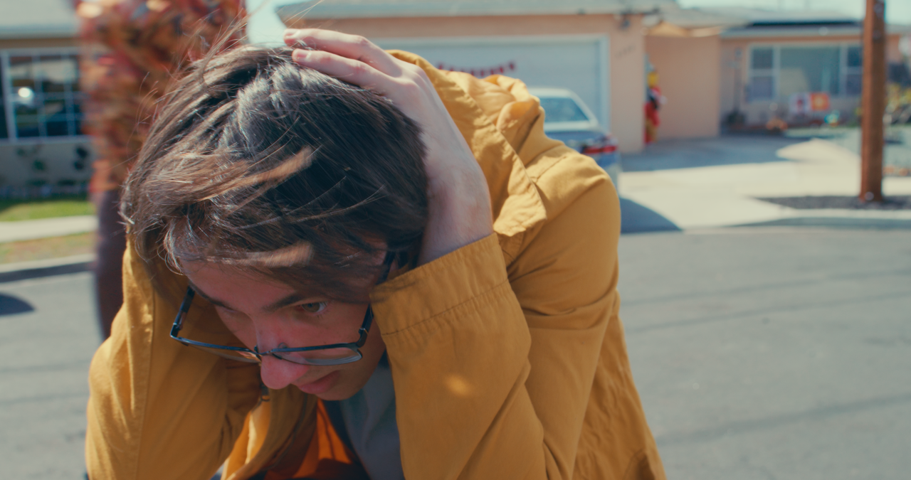 Young man wearing glasses in a yellow jacket, leaning forward with his hands on his head, outdoors on a sunny day with buildings and cars in the background.