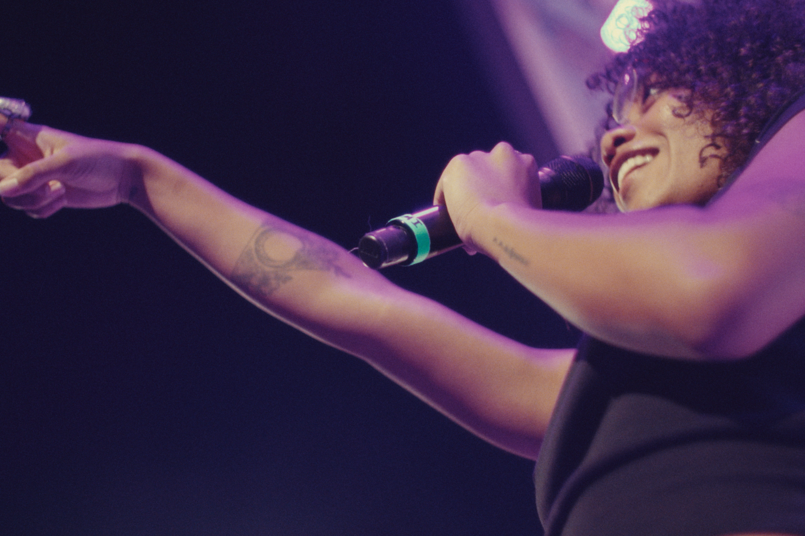 A woman singing into a microphone on stage, pointing with her right hand, with purple stage lighting.