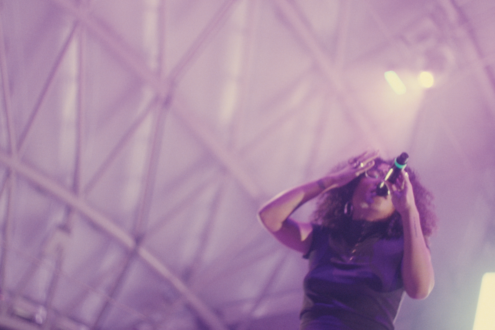 A woman with curly hair wearing glasses and a black shirt, singing into a microphone on stage, with purple stage lighting and a metal truss background.