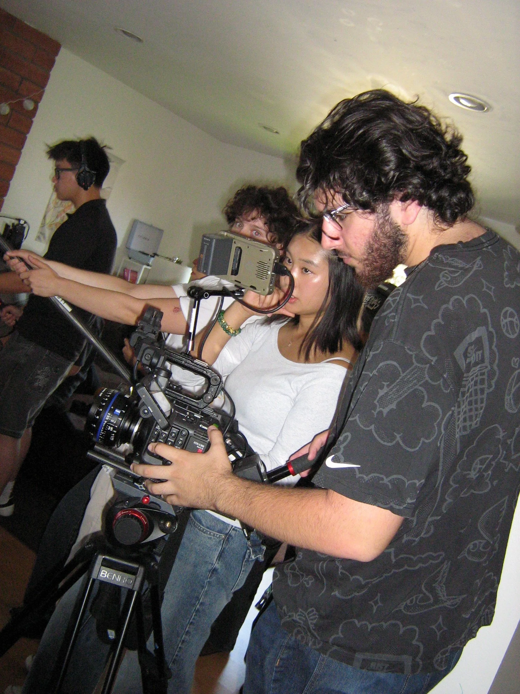 Group of people operating camera and filming equipment, one person holding a professional camera mounted on a tripod, others using electronic devices, indoors with white walls and ceiling lights.