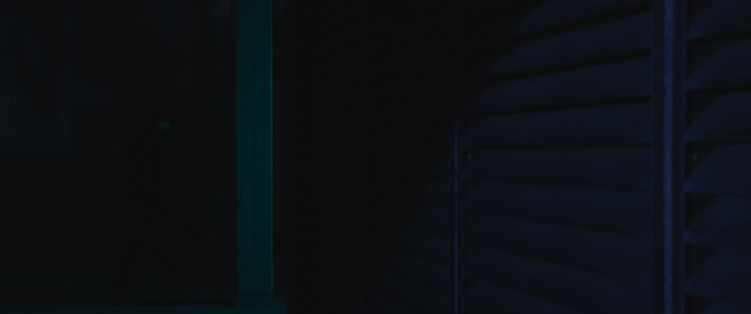Dark indoor space with a faintly visible blue structure on the right and a vertical green-lit element on the left.