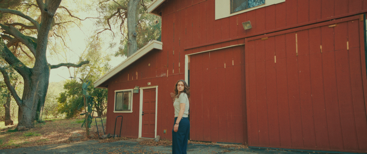 A woman standing outside a red barn-style building in a wooded area during daytime