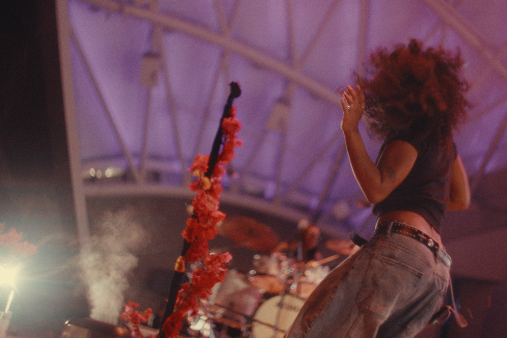 A woman with curly red hair dancing at a concert, wearing a black sleeveless top and blue jeans, with a floral decoration on a microphone stand and a drum set in the background, under purple and pink stage lighting.