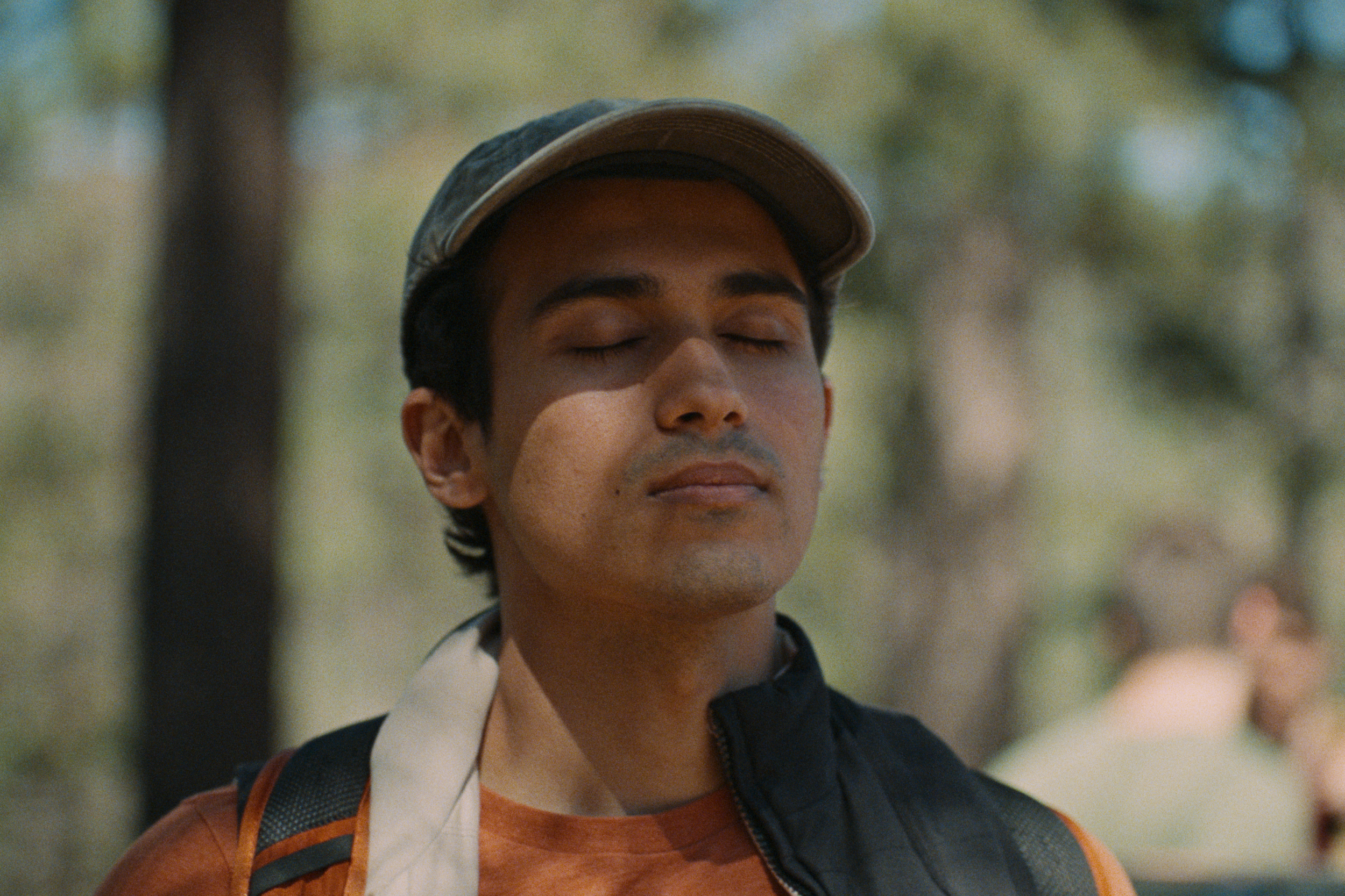 Close-up of a young man wearing a cap, eyes closed, with a backpack over one shoulder, outdoors in a park with blurred trees in the background.