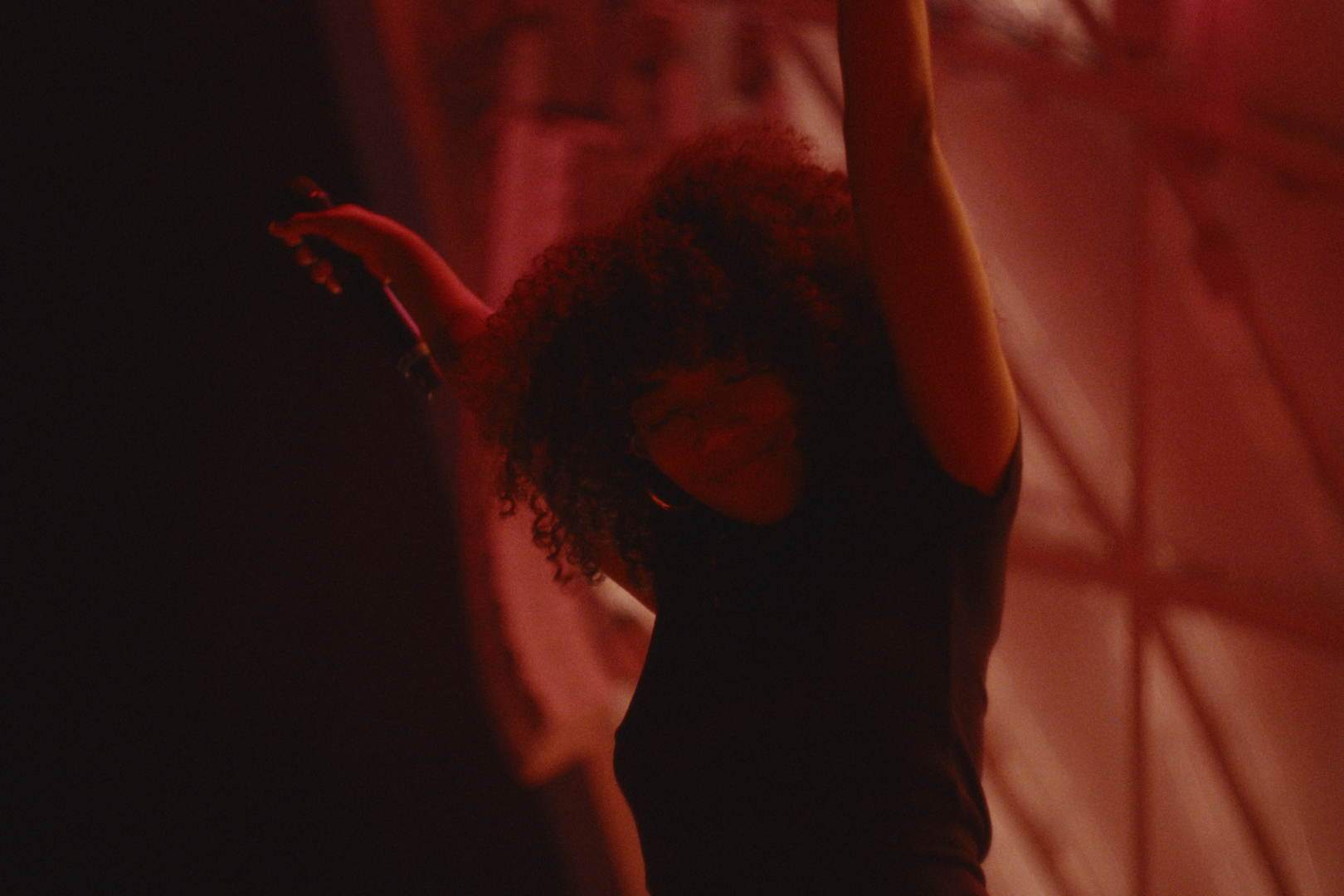 A woman with curly hair raising her arm in a dimly lit room with red and black lighting.