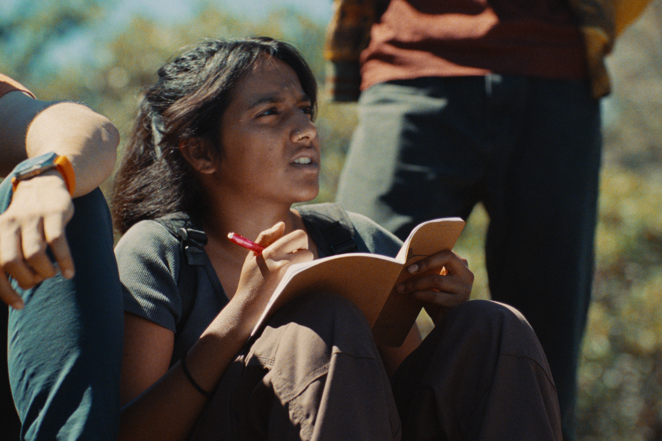 Young woman with curly hair and a nose piercing, sitting outdoors, holding a notebook and pen, surrounded by other people.