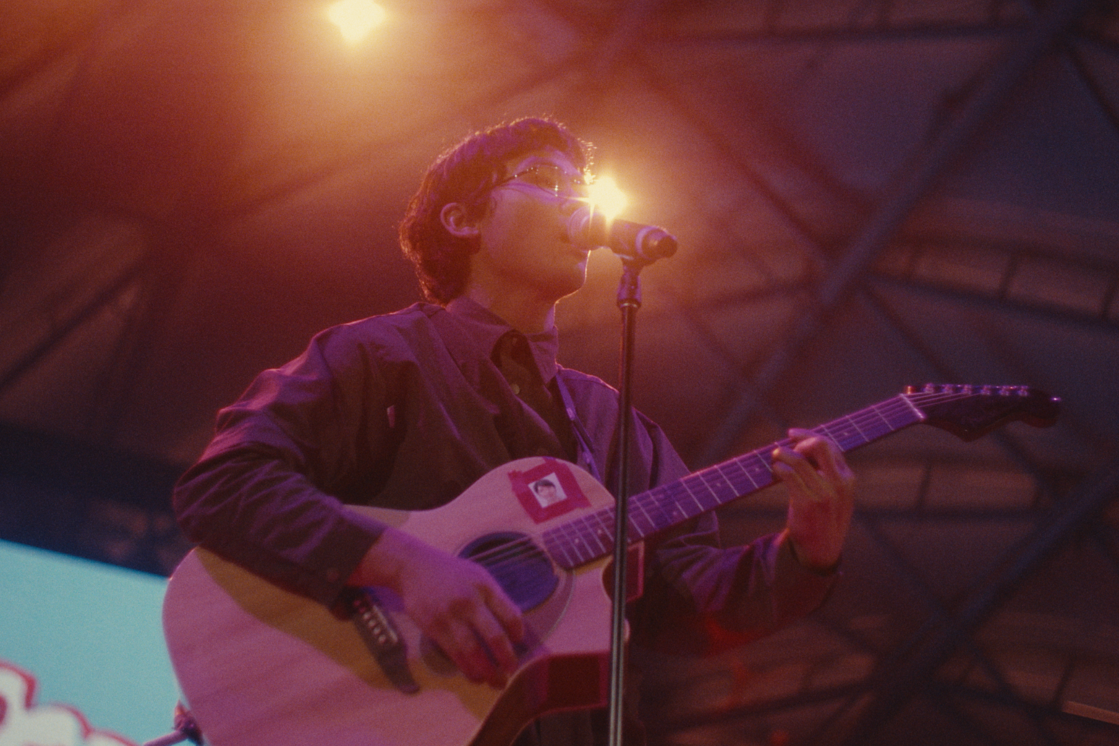 A person with dark hair and glasses playing an acoustic guitar and singing into a microphone under stage lighting.