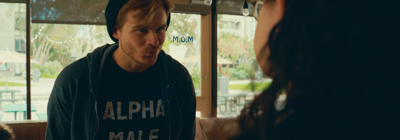 A young man with reddish hair and a beard talking to a woman inside a café, with an outdoor seating area visible through the window.
