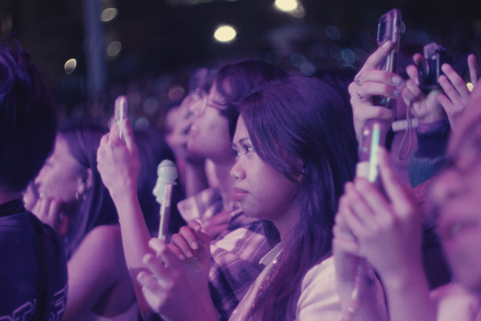A young woman with long dark hair holding a smartphone at a concert or event, surrounded by other people also taking photos or videos, with dim colorful lights in the background.