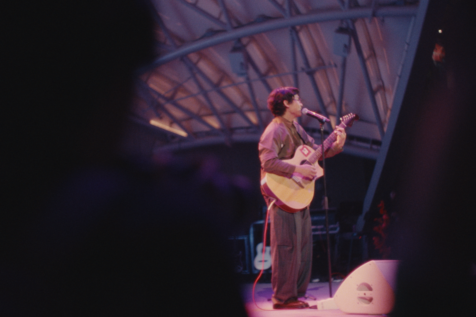 A person with curly hair, glasses, a brown jacket, and striped pants playing an acoustic guitar on stage under a high, metal-arched roof with stage lights.