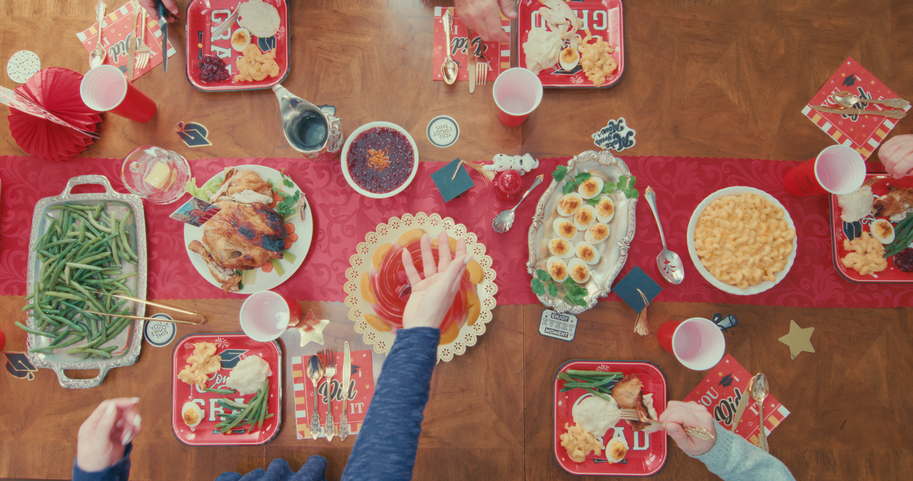 A festive holiday dining table set with plates of green beans, chicken, macaroni and cheese, deviled eggs, and desserts like banana pudding and cake. There are red cups, decorative napkins, and holiday-themed decorations.