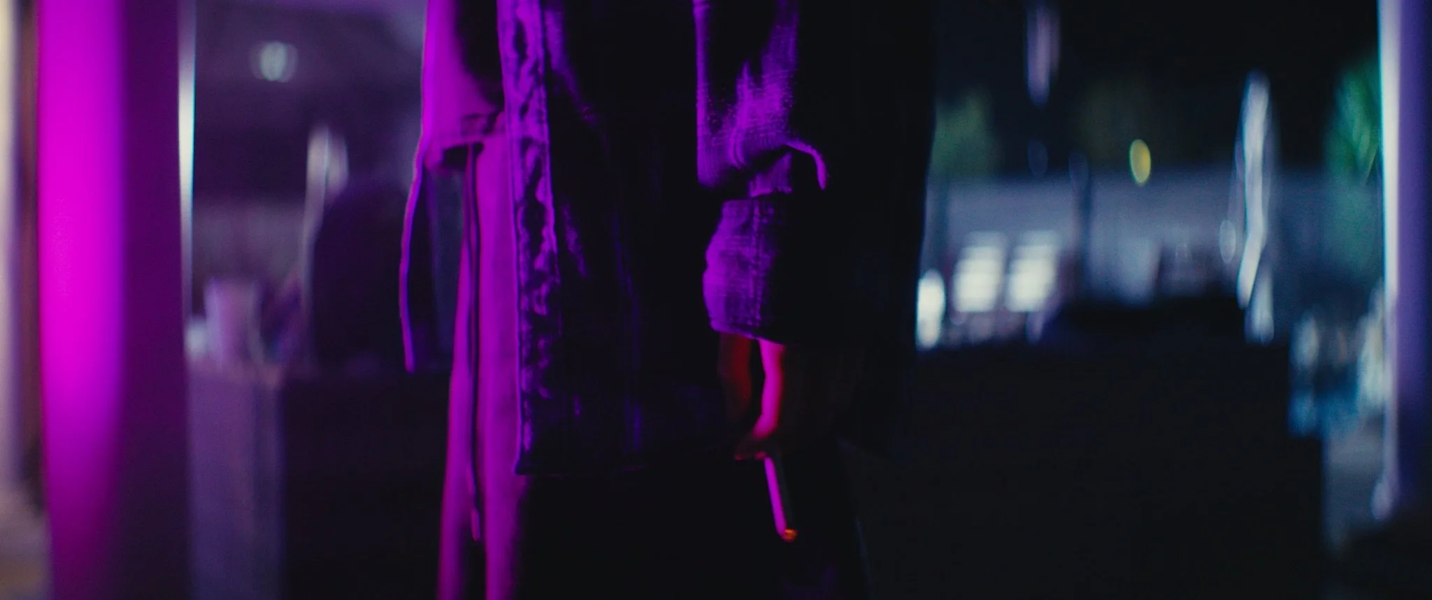 A person wearing dark jeans and a long coat standing in a dimly lit space with purple and blue neon lighting, holding a glowing stick.