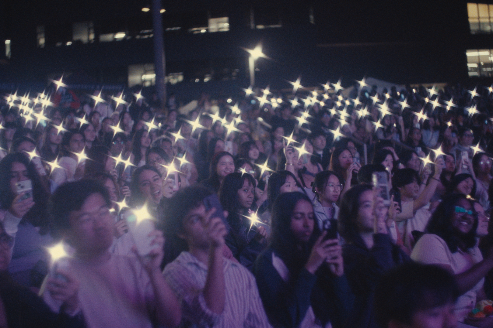 Crowd of people at an outdoor concert or event at night, many holding smartphones with the screens or flashes illuminated.