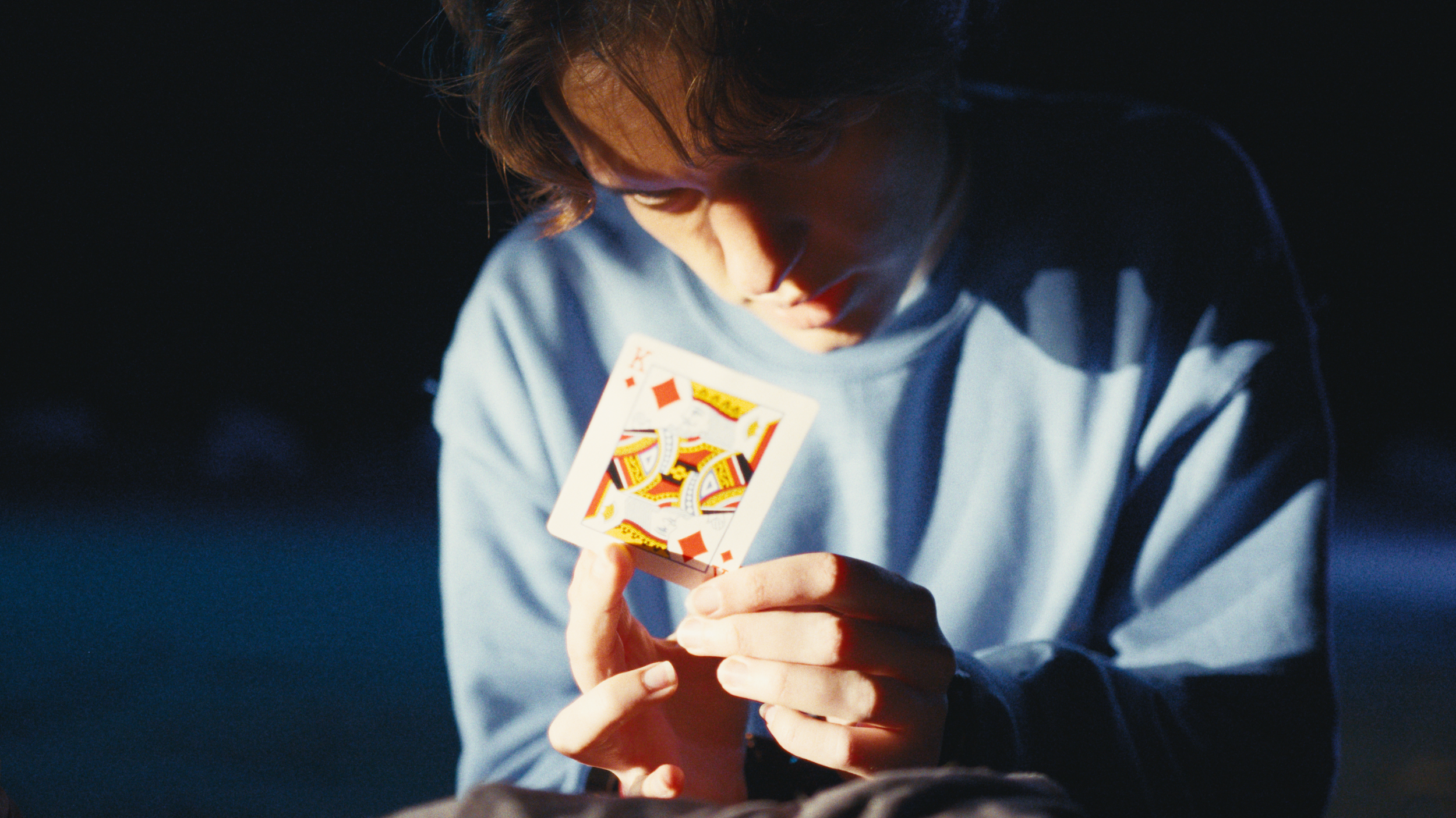 A person with medium-length hair looking down while holding a playing card, the Queen of Hearts, near their face.