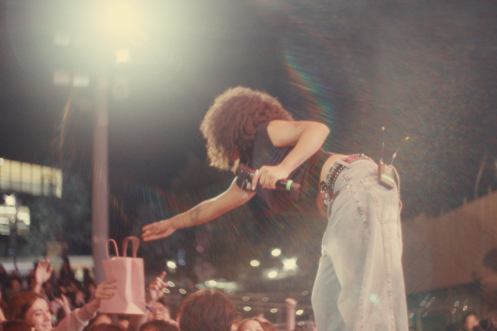 A female performer leaning forward on stage, holding a microphone, engaging with the audience at a concert or event, with crowd members reaching out.