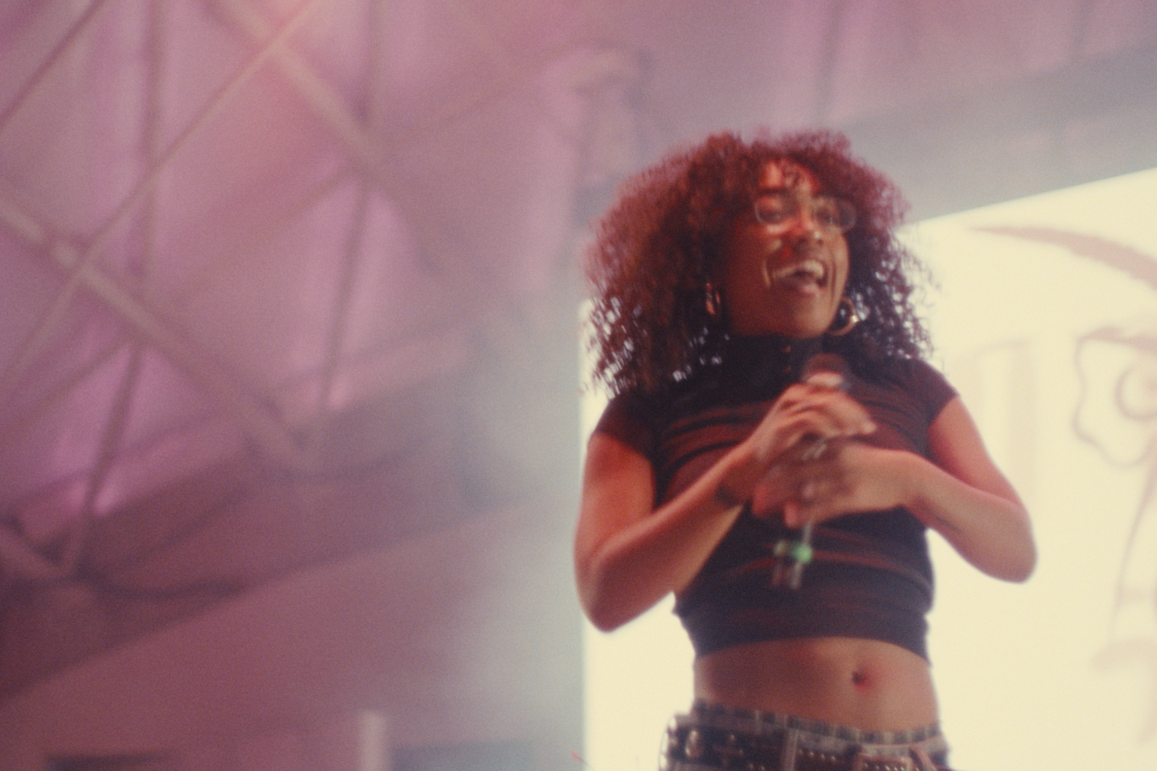 A woman with curly red hair, glasses, and hoop earrings, smiling and holding a microphone on stage with colorful lighting and screen in background.