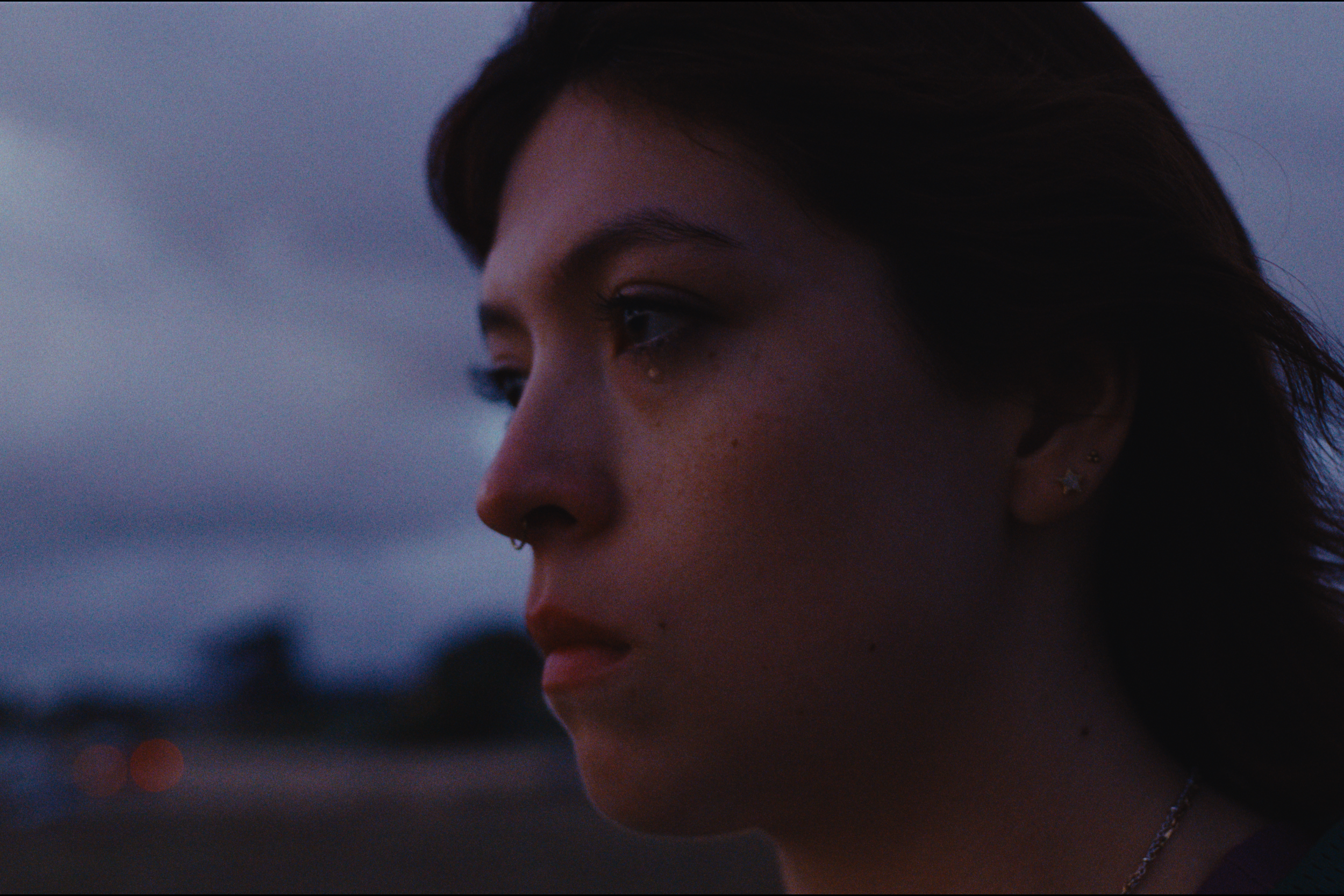 Close-up of a young woman's face with dark hair, earrings, and a serious expression, during dusk or evening.