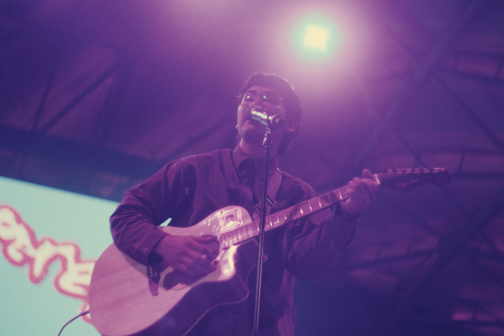 A man wearing glasses and a dark shirt is performing on stage, playing guitar and singing into a microphone, highlighted by purple stage lighting.
