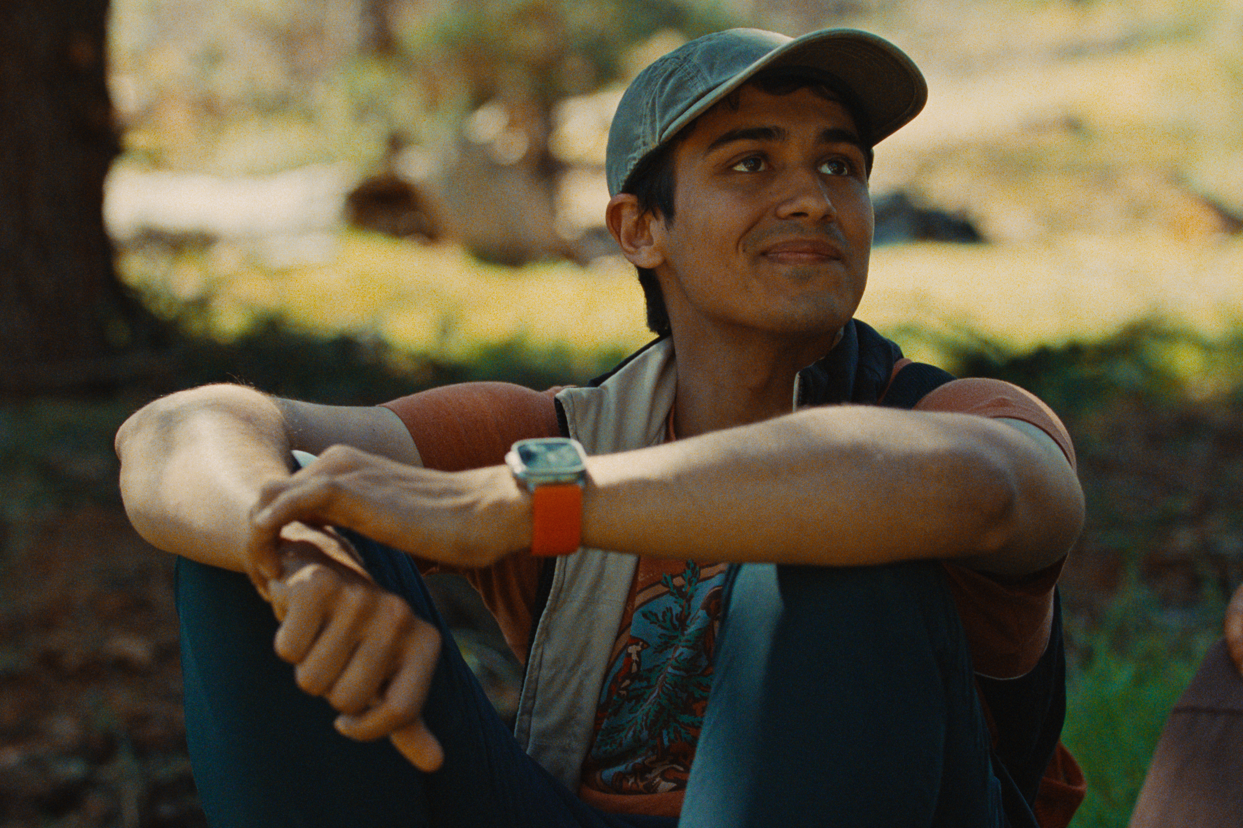 A young man with a tan complexion wearing a baseball cap and a colorful T-shirt sitting outdoors in a natural setting, possibly in a park or forest, with trees and foliage in the background.