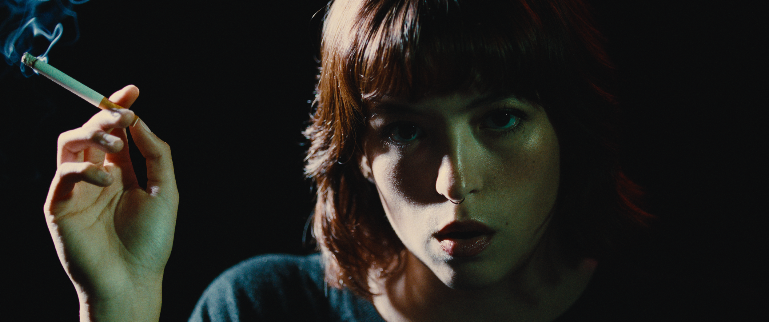 A woman with short red hair and dark lipstick holds a cigarette in her hand, with a dark background.