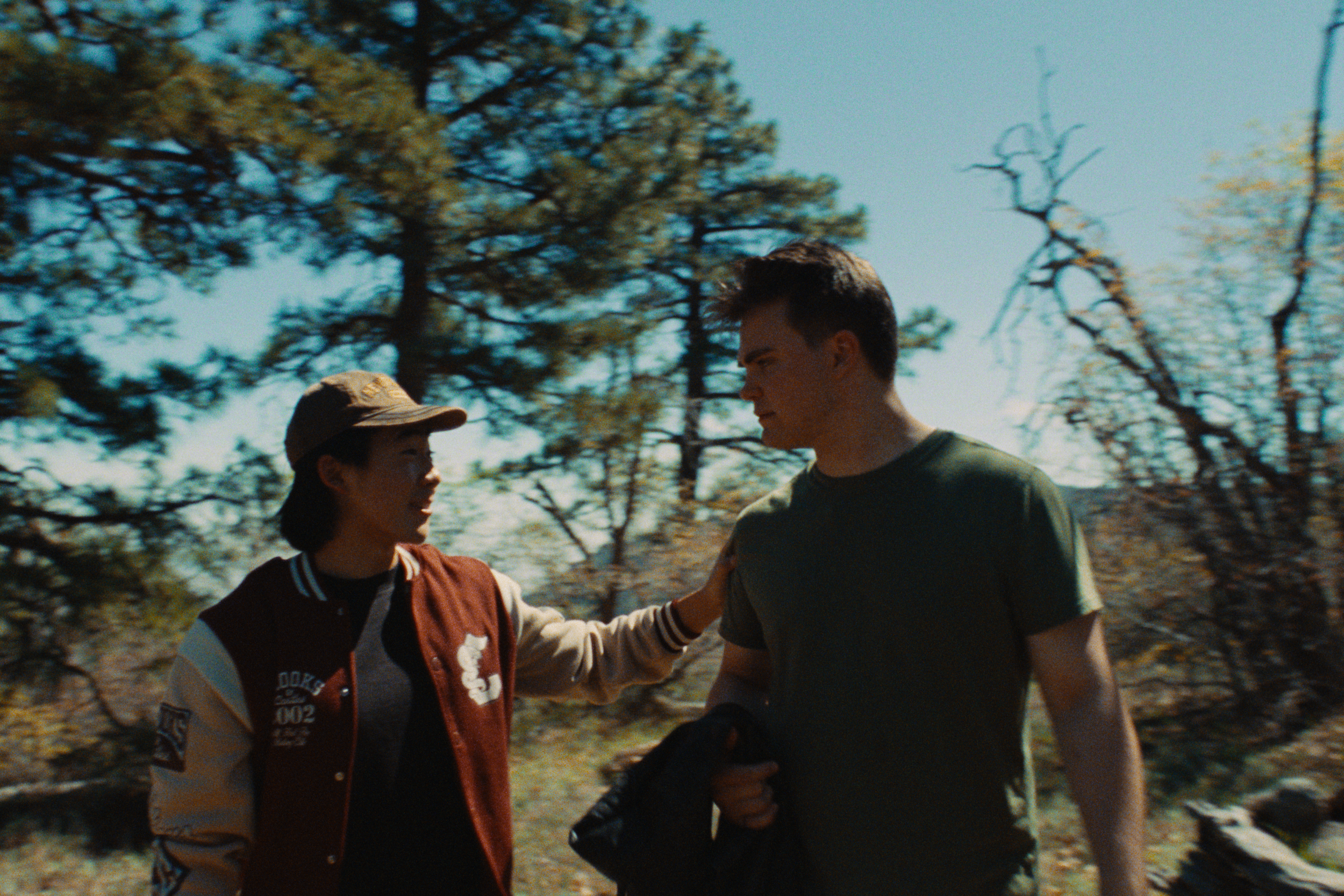 Two people, a woman and a man, are having a conversation outdoors, with trees in the background and mid-day sunlight.