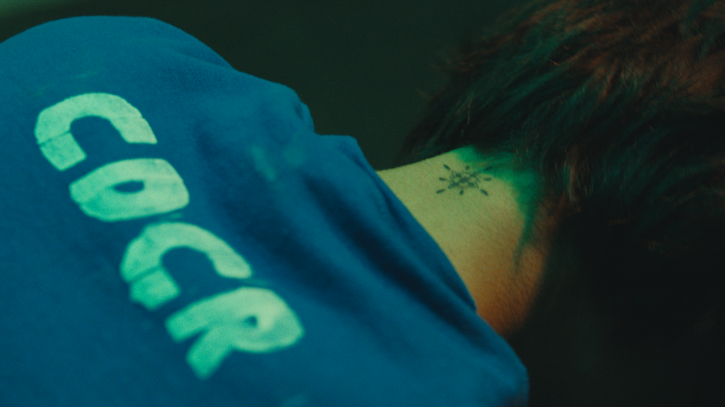 Close-up of a person with a tattoo on their neck, wearing a blue shirt with the word 'SOCCER' partially visible.
