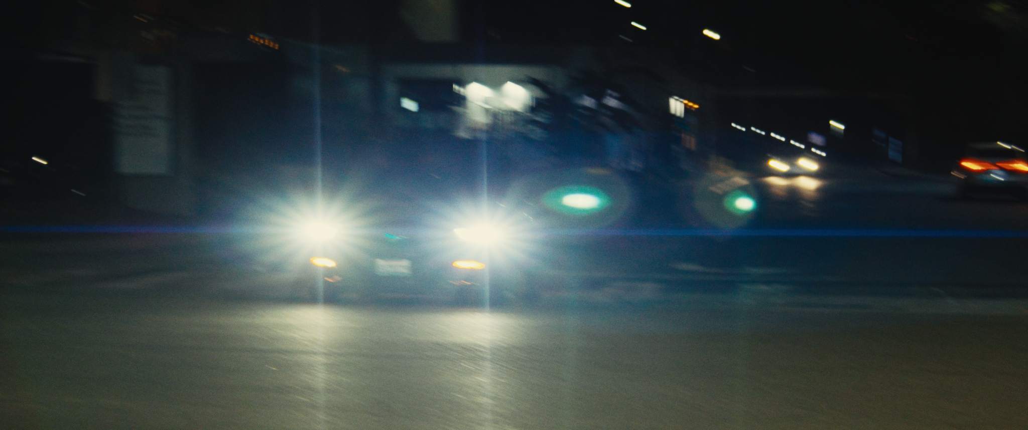 Nighttime scene of a city street with cars driving by, their headlights and taillights visible, and some buildings and streetlights in the background.