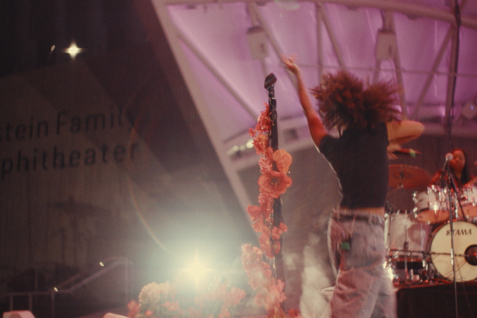 A performer with curly hair dancing on stage with a microphone in hand, wearing a black crop top and jeans, surrounded by musical instruments and stage decorations, with stage lights creating a vibrant atmosphere.
