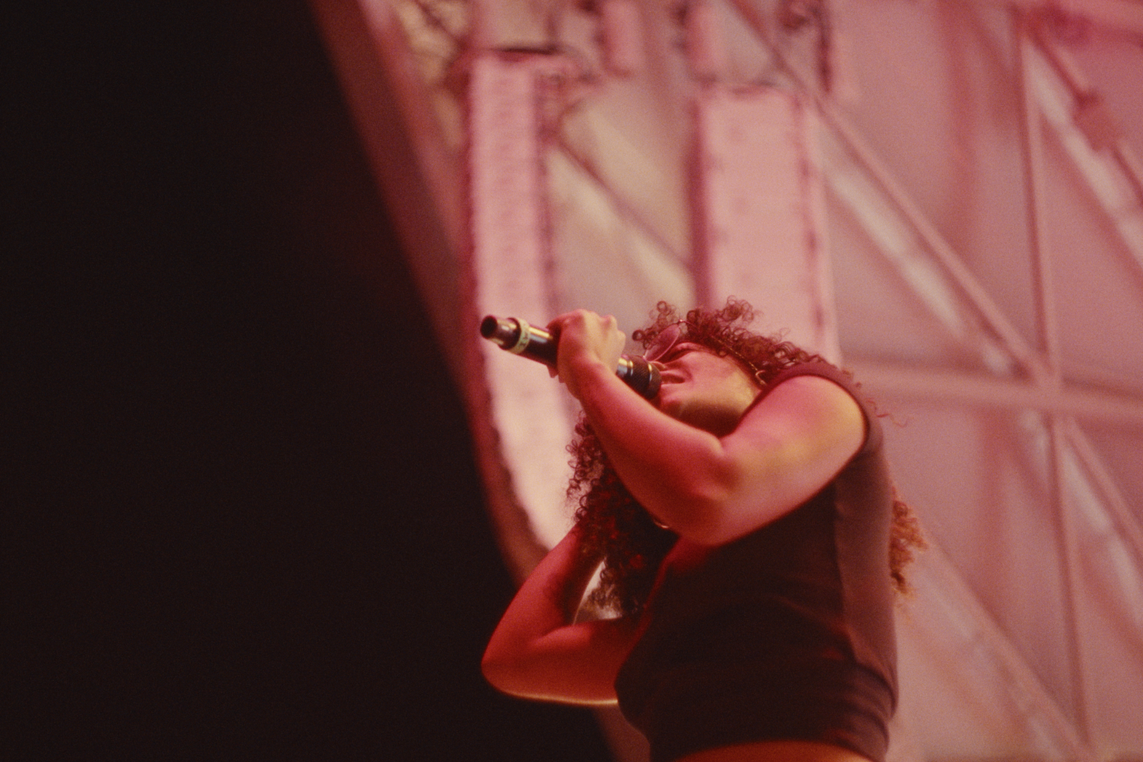 A woman with curly hair sings into a microphone on stage with a metal structure in the background and reddish lighting.