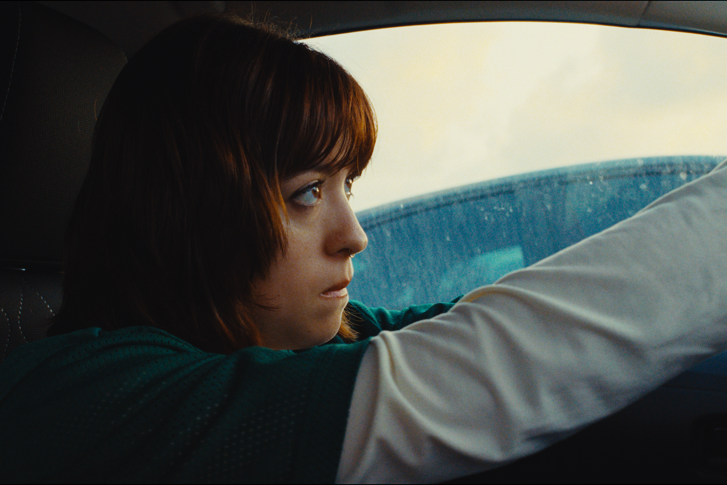 A young woman with red hair and blue eyes sitting in the passenger seat of a car, looking intently out the window.