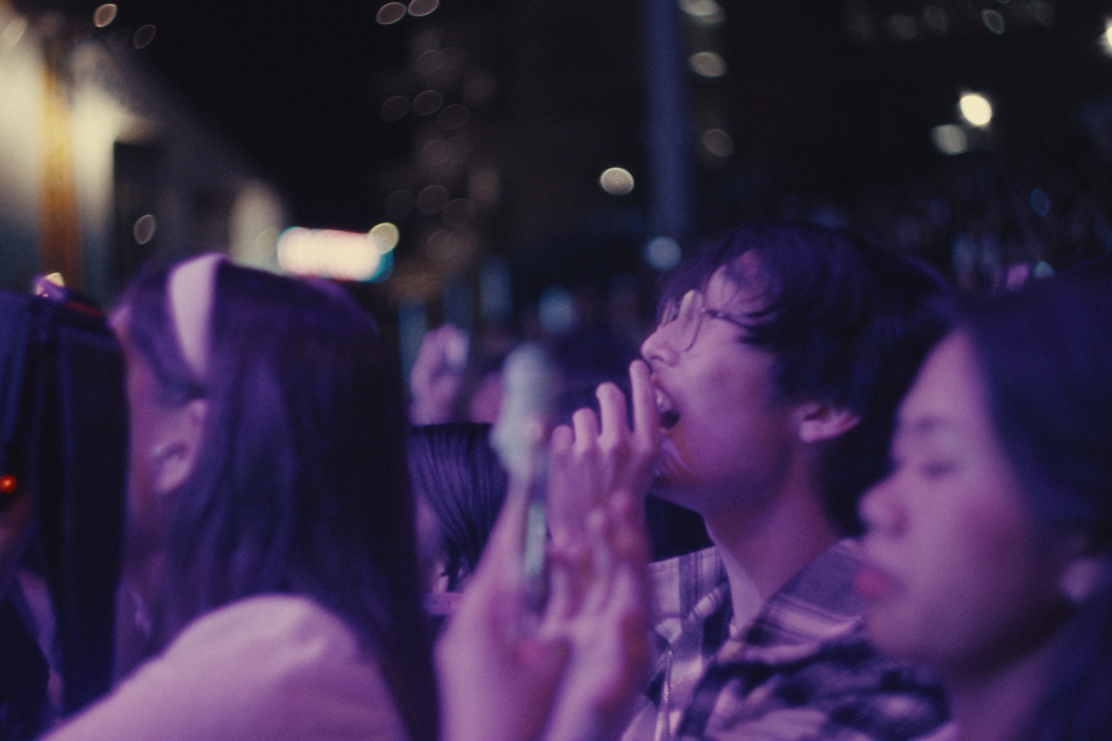 People in a dimly lit, nighttime setting with colorful purple and blue lighting, some individuals with their hands raised, engaging in a lively event or concert.