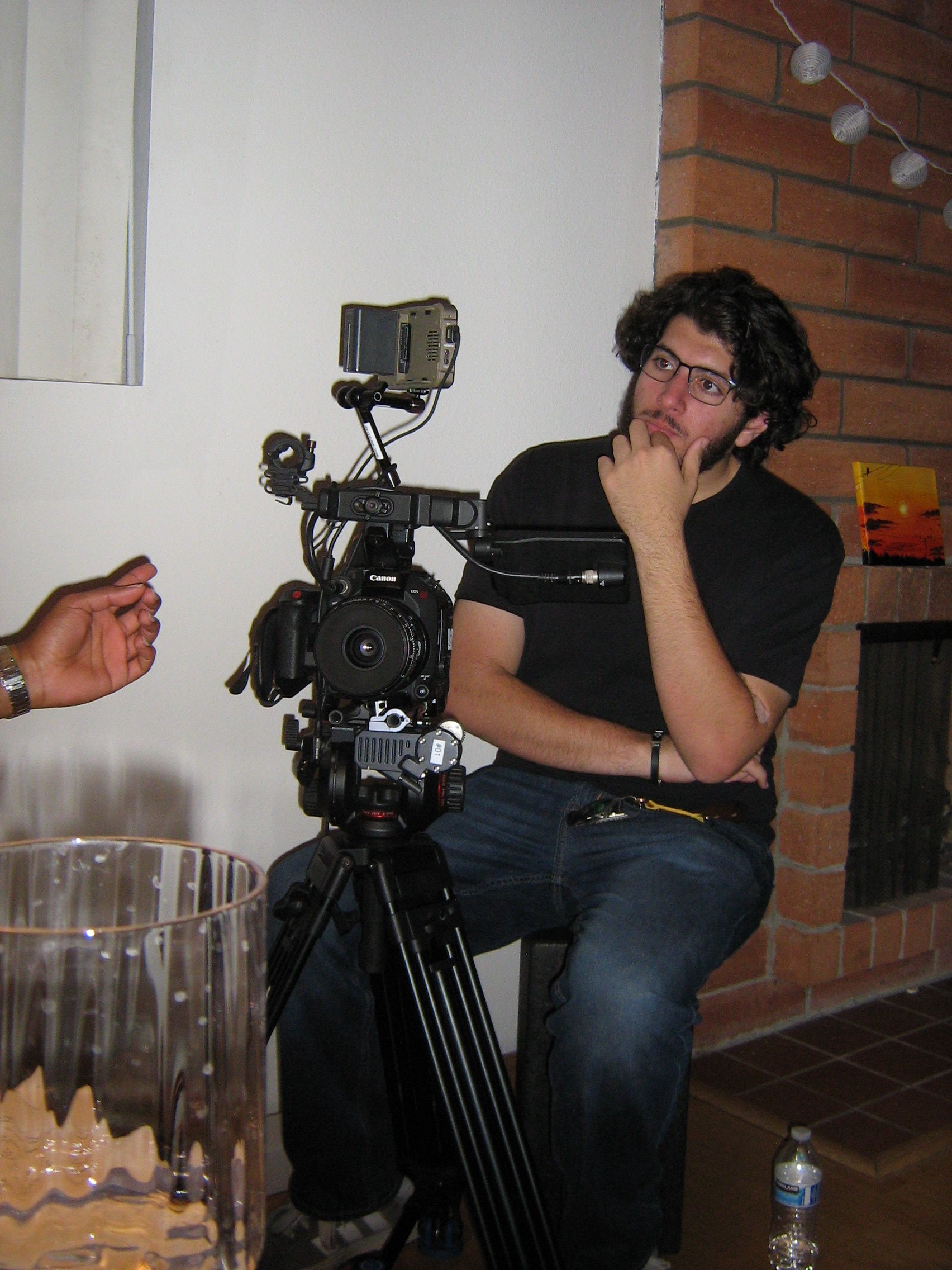 A man with glasses and curly hair sits with his hand on his chin, looking thoughtful, next to a professional camera mounted on a tripod. There is a glass and a water bottle on the table in front of him. The room has a brick wall and a white wall with some string lights hanging.